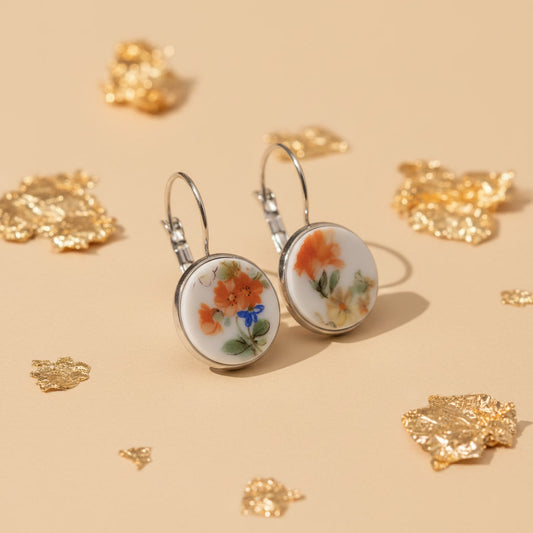 Produktaufnahme , seitlich fotografiert, von zwei runden Upcycling-Ohrhängern aus weißem Porzellan mit einem handgemalten Blumenmotiv in Orange und Gelb, präsentiert auf einem beigefarbenen Untergrund mit dekorativen Goldflocken.