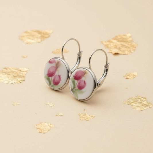 Nahaufnahme von zwei handgefertigten Upcycling-Ohrhängern aus weißem Porzellan, seitlich fotografiert, mit einem filigranen Blumenmotiv in dunklem Pink und Grün, gefasst in silberfarbenen Edelstahl auf einem beigefarbenen Hintergrund mit dekorativen Goldflocken.