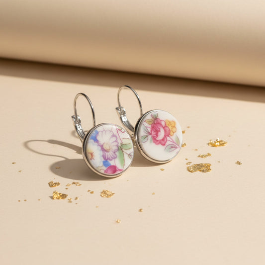 Seitenansicht der Varietas Porzellan-Ohrringe von Edelatelier, die die hochwertigen Klappbrisuren aus silberfarbenem Edelstahl zeigen. Die flachen, handgeschliffenen Porzellan-Inlays mit ihren farbenfrohen Blumenmustern sind deutlich erkennbar. Die Ohrhänger stehen auf einer beigefarbenen Fläche mit Goldakzenten, was das leichte und elegante Design unterstreicht.