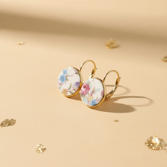 Nahaufnahme der Millefleurs-Ohrringe, die leicht versetzt auf einem beigen Untergrund liegen. Das detaillierte florale Muster und die glänzenden, goldfarbenen Klappbrisuren aus Edelstahl sind deutlich sichtbar und unterstreichen die hochwertige Verarbeitung.