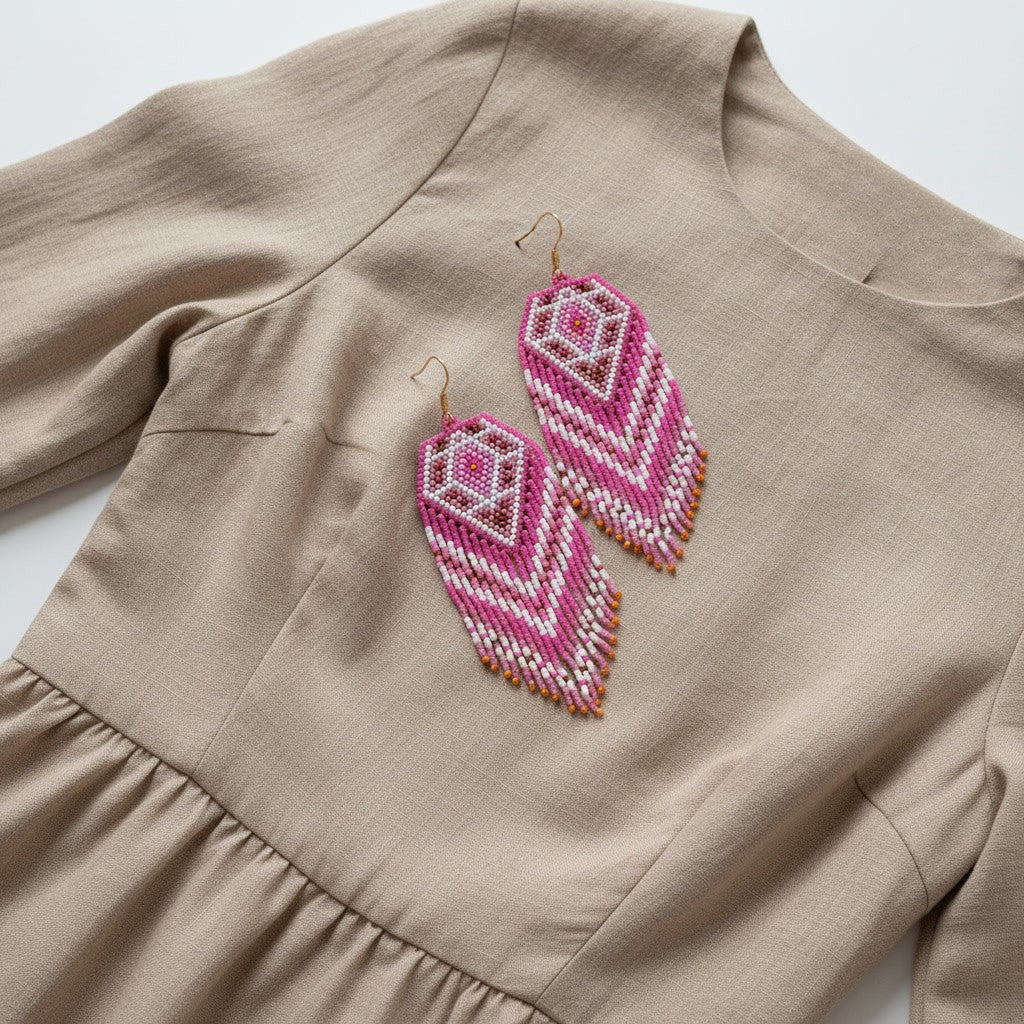 Ein professionelles Flatlay-Foto auf hellem Hintergrund zeigt ein beigefarbenes Kleid aus Leinenstoff, auf dessen Brustbereich ein Paar handgefertigte Perlenohrringe von EDELATELIER. platziert ist. Die opulenten Statement-Ohrringe sind in einem geometrischen Rautenmuster gewebt und bestechen durch ein harmonisches Farbspiel aus kräftigem Pink, zartem Rosa und reinem Weiß. Die langen Fransen an der Unterseite verleihen dem Design eine fließende Dynamik.