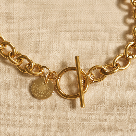 Detailaufnahme der Catena Luce Halskette aus 18 Karat PVD-vergoldetem Edelstahl mit T-Verschluss und EDELATELIER-Logoplättchen auf warmem hellbeigem Leinen.