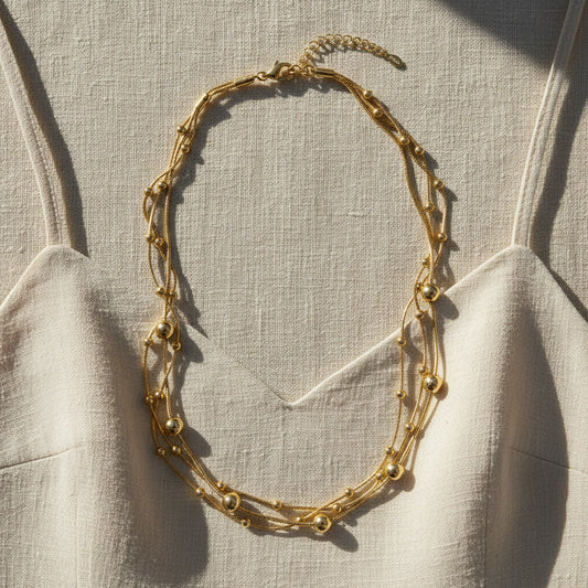 Mehrreihige goldfarbene Halskette mit Kugeldetails auf hellem Leinen-Top im warmen Sonnenlicht.