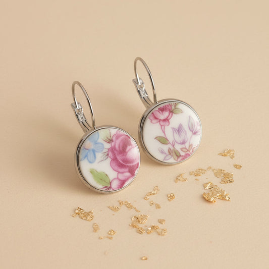 Seitenansicht der Floris Porzellan-Ohrringe von Edelatelier, die die hochwertigen Klappbrisuren aus Edelstahl zeigen. Die runden Inlays aus Upcycling-Porzellan präsentieren eine abwechslungsreiche Blütenpracht mit einer großen pinkfarbenen Rose links und filigranen violetten sowie hellblauen Blüten rechts. Das Paar ist auf einem beigefarbenen Hintergrund mit dekorativen Goldflocken und sanfter Schattenbildung arrangiert.