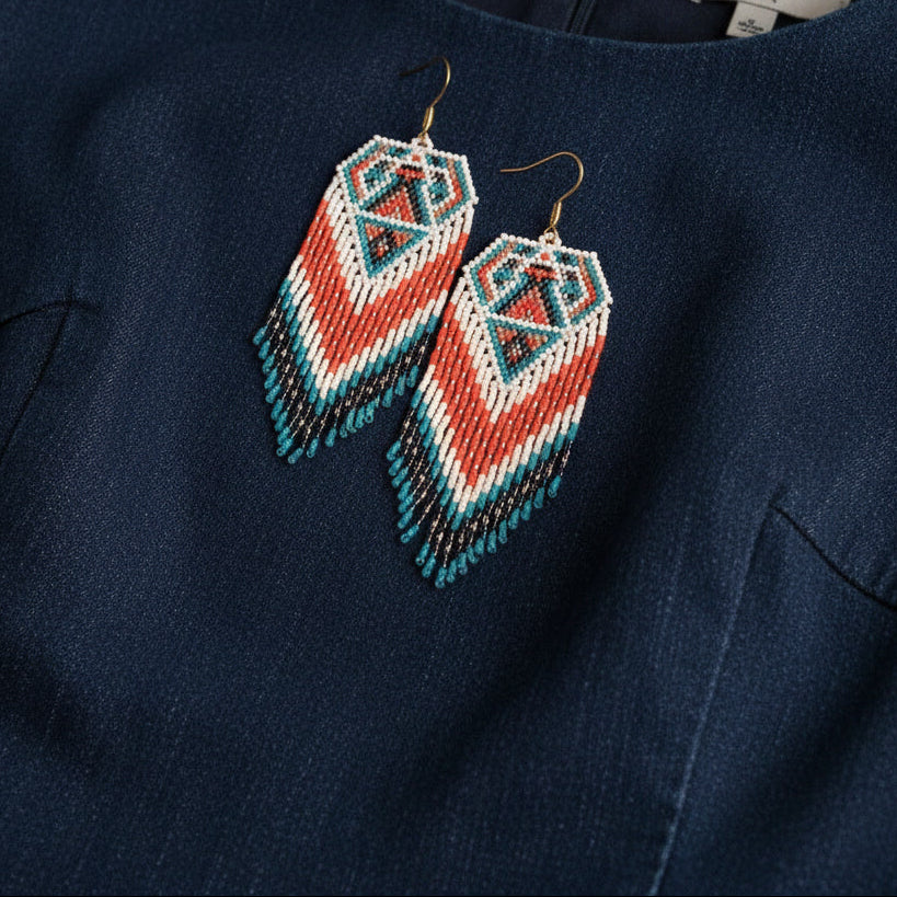 Eine Nahaufnahme handgefertigter Perlenohrringe mit einem geometrischen Muster aus Türkis, Grün, Orange und Gelb, die auf einem schönen schwarzen Jeanskleid liegen. Ein weicher Lichtstrahl mit gestreiften Schatten fällt schräg über die Szene.