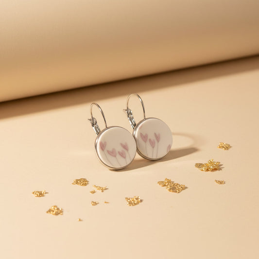 Handgefertigte Porzellanohrringe „Flosculus“ von EDELATELIER mit zartrosa Tulpenmotiven auf weißem Porzellan, eingefasst in silberfarbene Rundfassungen, präsentiert auf beigem Hintergrund mit Goldakzenten.
