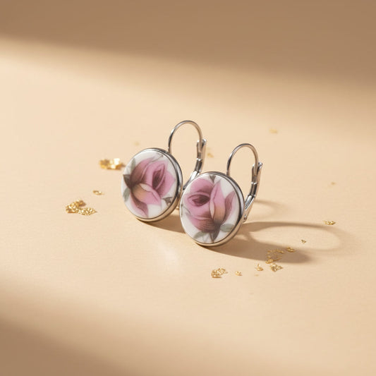 Detailaufnahme der handgefertigten Porzellanohrringe Rosa Splendida von EDELATELIER aus einer schrägen Seitenperspektive. Die beiden runden Ohrhänger stehen aufrecht auf einem beigefarbenen Untergrund, der mit dekorativen Goldflocken bestreut ist. Diese Ansicht betont die flache, elegante Form der Porzellanscherben und die hochwertige Einfassung in silberfarbene Fassungen. Die silbernen Ohrhaken (Brisuren) sind deutlich in ihrer geschwungenen Form erkennbar. Das Licht fällt von rechts ein und erzeugt einen