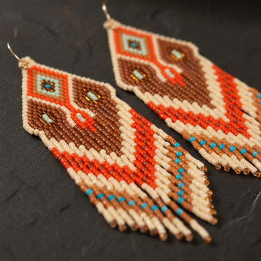 Detaillierte Makroaufnahme des handgewebten Rautenmusters der Statement-Ohrringe Sabbia Dorata von EDELATELIER. Die Komposition aus matten Miyuki-Perlen in Terrakotta, kräftigem Orange und Meeresblau mit goldenen Akzenten zeigt die hochwertige Handwerkskunst und feine Struktur auf dunklem Naturschiefer.