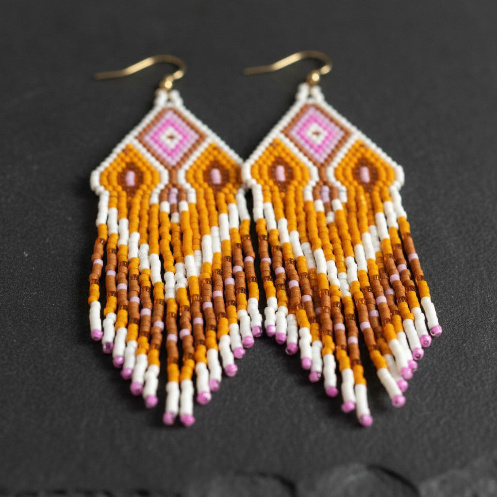 Ein Paar handgefertigte Perlenohrringe von EDELATELIER auf einem dunklen, schieferartigen Untergrund. Das Design besticht durch eine helle Farbkombination: Ein weißes Rautenmuster rahmt eine zentrale Raute in kräftigem Pink und zartem Rosé ein. Darunter fließen lange Fransen in warmem Ockergelb, Weiß und Braun, die an den Spitzen mit pinkfarbenen Akzenten abschließen. Die Ohrringe sind an goldfarbenen Haken befestigt und zeigen die präzise Webtechnik der Miyuki-Perlen.