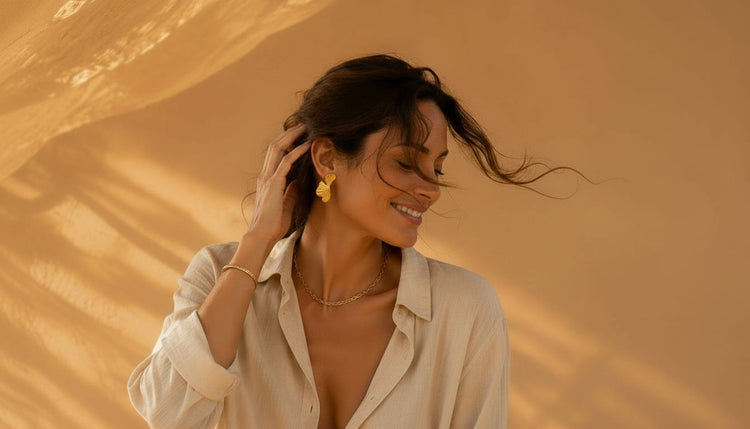 Model mit goldfarbenem Edelstahlschmuck vor warmbeigem Hintergrund im Sonnenlicht.