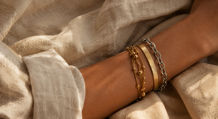 Brauner Arm mit gold- und silberfarbenen Edelstahl-Armbändern im Layering-Look auf beigem Leinenstoff.