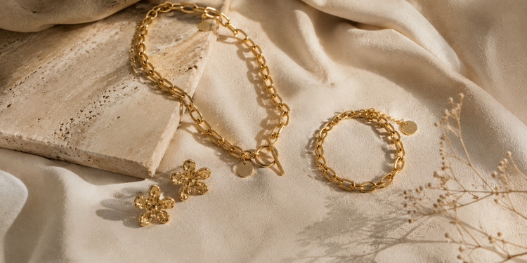 Goldfarbene Edelstahl Schmuckstücke von EDELATELIER auf beigem Stoff mit Travertinplatte im warmen Sonnenlicht.