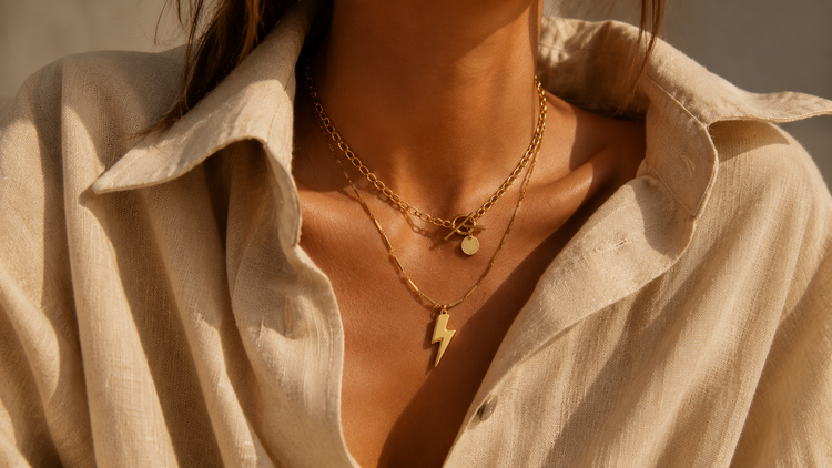 Goldfarbene Halsketten im Layering-Look auf brauner Haut mit heller Leinenbluse im Sonnenlicht.