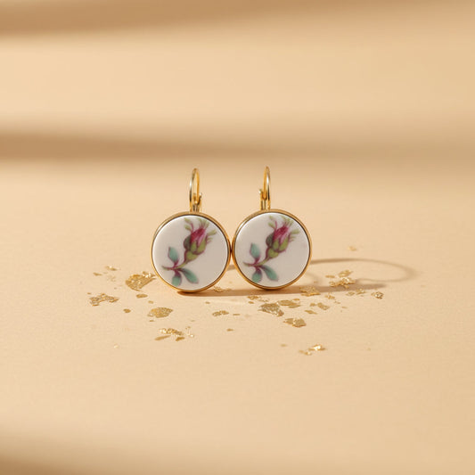 Ein Paar handgefertigte Porzellanohrringe "Alba Rosa" von EDELATELIER. auf einem hellen, beigen Hintergrund mit dezenten Goldpartikeln. Die runden Ohrhänger zeigen eine zarte, pinkfarbene Rosenknospe mit grünen Blättern auf reinweißem Porzellan, sicher eingefasst in eine glänzende, goldfarbene Edelstahlfassung.