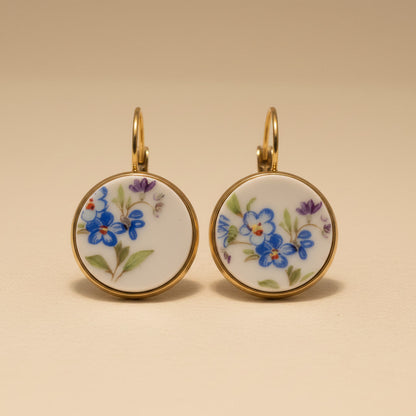 Elegante Porzellanohrringe mit zarten blauen Vergissmeinnicht-Motiven und violetten Akzenten in einer hochwertigen Gold-Optik vor einem beigen Hintergrund - frontal fotografiert. 