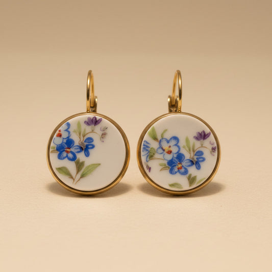 Elegante Porzellanohrringe mit zarten blauen Vergissmeinnicht-Motiven und violetten Akzenten in einer hochwertigen Gold-Optik vor einem beigen Hintergrund - frontal fotografiert. 