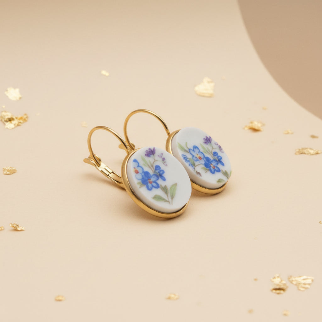 Elegante Porzellanohrringe mit zarten blauen Vergissmeinnicht-Motiven und violetten Akzenten in einer hochwertigen Gold-Optik vor einem beigen Hintergrund mit Blattgold als Dekor, seitlich fotografiert. 