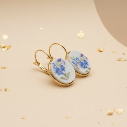 Elegante Porzellanohrringe mit zarten blauen Vergissmeinnicht-Motiven und violetten Akzenten in einer hochwertigen Gold-Optik vor einem beigen Hintergrund mit Blattgold als Dekor, seitlich fotografiert. 