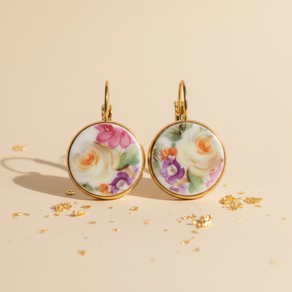 Frontansicht der Aurelia Porzellan-Ohrringe von Edelatelier, welche die hochwertigen Klappbrisuren aus goldfarbenem Edelstahl zeigen. Die runden Inlays aus Upcycling-Porzellan präsentieren ein prachtvolles Rosenmotiv, bei dem sanfte Gelbtöne im Fokus stehen, ergänzt durch Akzente in Pink und Violett. Das Paar ist auf einem beigefarbenen Hintergrund mit dekorativen Goldflocken und warmem Lichteinfall arrangiert.