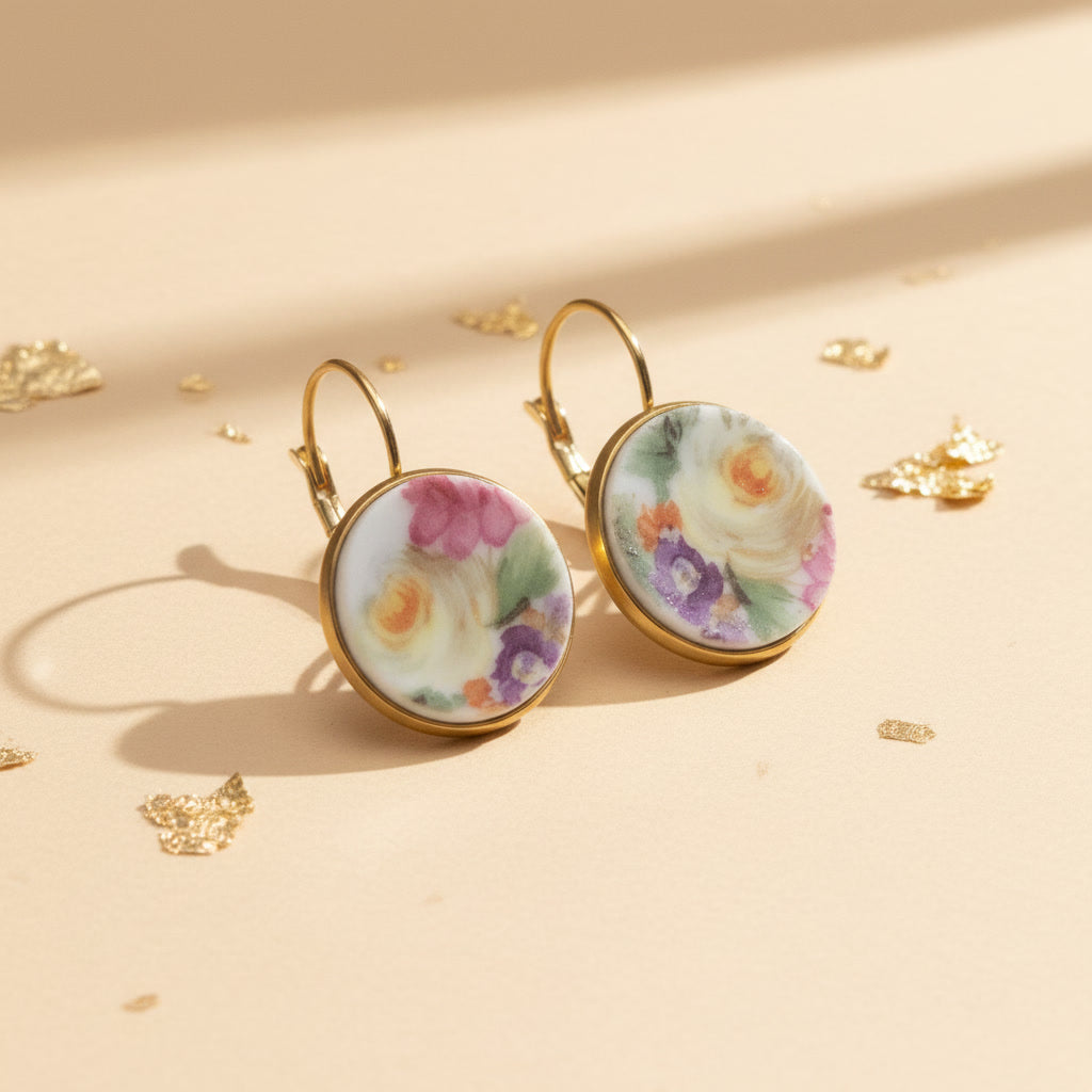 Seitenansicht der Aurelia Porzellan-Ohrringe von Edelatelier, welche die hochwertigen Klappbrisuren aus goldfarbenem Edelstahl zeigen. Die runden Inlays aus Upcycling-Porzellan präsentieren ein prachtvolles Rosenmotiv, bei dem sanfte Gelbtöne im Fokus stehen, ergänzt durch Akzente in Pink und Violett. Das Paar ist auf einem beigefarbenen Hintergrund mit dekorativen Goldflocken und warmem Lichteinfall arrangiert.
