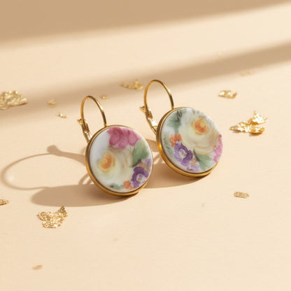 Seitenansicht der Aurelia Porzellan-Ohrringe von Edelatelier, welche die hochwertigen Klappbrisuren aus goldfarbenem Edelstahl zeigen. Die runden Inlays aus Upcycling-Porzellan präsentieren ein prachtvolles Rosenmotiv, bei dem sanfte Gelbtöne im Fokus stehen, ergänzt durch Akzente in Pink und Violett. Das Paar ist auf einem beigefarbenen Hintergrund mit dekorativen Goldflocken und warmem Lichteinfall arrangiert.
