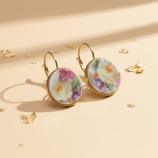 Seitenansicht der Aurelia Porzellan-Ohrringe von Edelatelier, welche die hochwertigen Klappbrisuren aus goldfarbenem Edelstahl zeigen. Die runden Inlays aus Upcycling-Porzellan präsentieren ein prachtvolles Rosenmotiv, bei dem sanfte Gelbtöne im Fokus stehen, ergänzt durch Akzente in Pink und Violett. Das Paar ist auf einem beigefarbenen Hintergrund mit dekorativen Goldflocken und warmem Lichteinfall arrangiert.