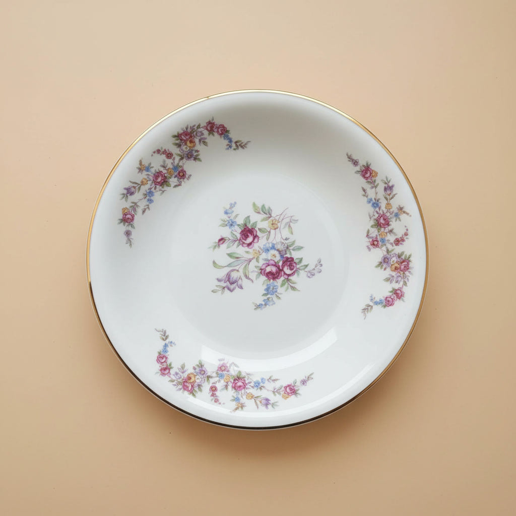 Ein runder Vintage-Teller aus weißem Porzellan mit Goldrand, der als Ursprung für die Schmuckstücke von Edelatelier dient. Das Dekor zeigt ein zentrales Bukett aus pinkfarbenen Rosen und blauen Vergissmeinnicht sowie umlaufende florale Ranken in Pastelltönen. Der Teller liegt auf einem ruhigen, beigen Hintergrund unter gleichmäßiger Beleuchtung.