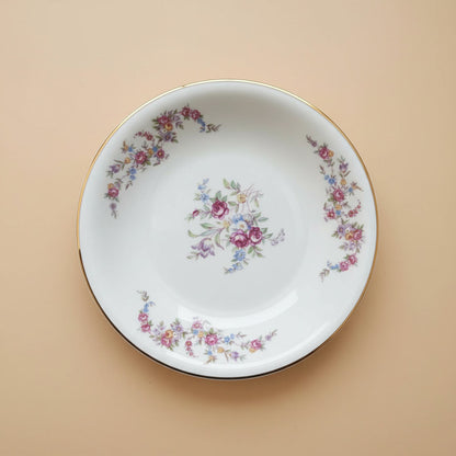 Ein runder Vintage-Teller aus weißem Porzellan mit Goldrand, der als Ursprung für die Schmuckstücke von Edelatelier dient. Das Dekor zeigt ein zentrales Bukett aus pinkfarbenen Rosen und blauen Vergissmeinnicht sowie umlaufende florale Ranken in Pastelltönen. Der Teller liegt auf einem ruhigen, beigen Hintergrund unter gleichmäßiger Beleuchtung.