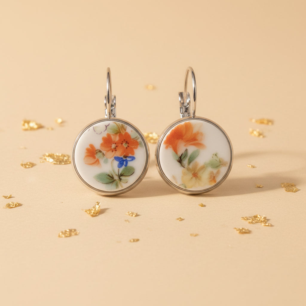 Nahaufnahme von zwei runden Porzellanohrringen mit handgeschliffenen Inlays aus weißem Vintage-Porzellan, verziert mit einem filigranen Blumenmuster in Orange, Gelb und einem kleinen blauen Akzent, eingefasst in silberglänzende Edelstahlfassungen auf einem beigefarbenen Hintergrund mit Goldflocken.
