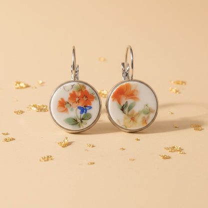 Nahaufnahme von zwei runden Porzellanohrringen mit handgeschliffenen Inlays aus weißem Vintage-Porzellan, verziert mit einem filigranen Blumenmuster in Orange, Gelb und einem kleinen blauen Akzent, eingefasst in silberglänzende Edelstahlfassungen auf einem beigefarbenen Hintergrund mit Goldflocken.