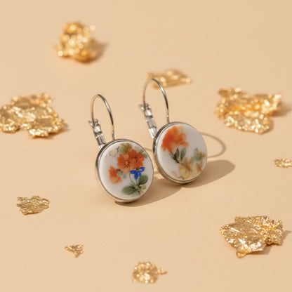 Produktaufnahme , seitlich fotografiert, von zwei runden Upcycling-Ohrhängern aus weißem Porzellan mit einem handgemalten Blumenmotiv in Orange und Gelb, präsentiert auf einem beigefarbenen Untergrund mit dekorativen Goldflocken.