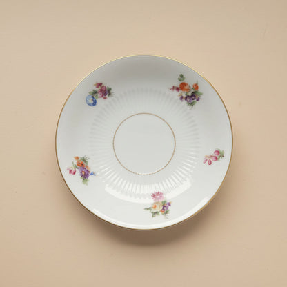 Ein antiker weißer Porzellanteller mit Goldrand und verschiedenen Streublumen-Motiven in Orange, Lila und Pink, der als Ausgangsmaterial für die handgefertigten Ohrringe vom Edelatelier dient.