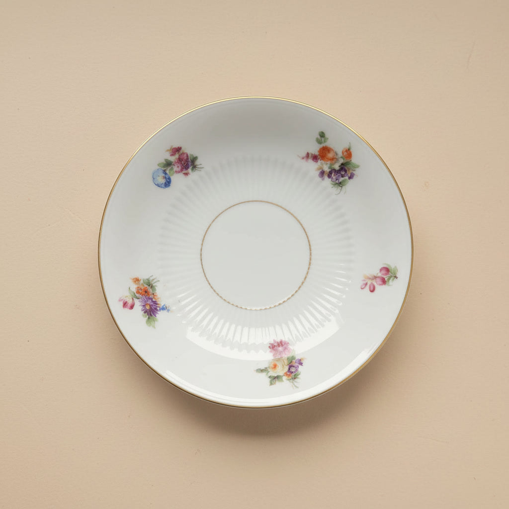 Ein weißer antiker Porzellanteller mit Goldrand und verschiedenen Streublumen-Motiven in Pink, Orange und Lila, der als Materialquelle für die handgefertigten Unikate von Edelatelier dient.