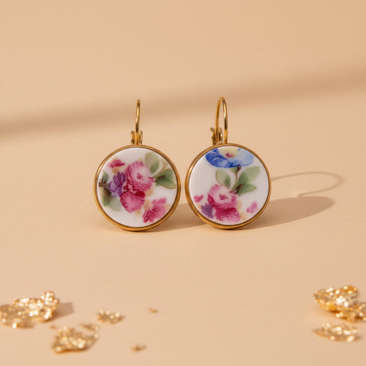 Nahaufnahme von zwei runden Upcycling-Ohrhängern aus weißem Porzellan mit einem reichhaltigen, bunten Floribunda-Blumenstrauß-Motiv in Pink, Blau und Lila, eingefasst in goldfarbenem Edelstahl auf beigem Hintergrund mit Blattgold auf dem Untergrund.