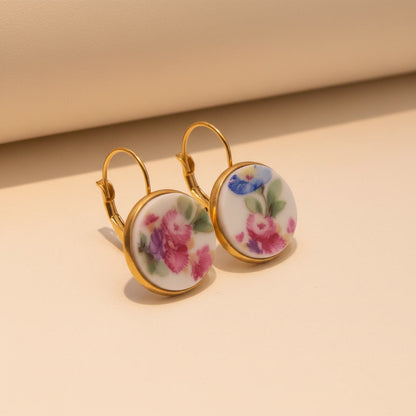 Nahaufnahme von zwei runden Upcycling-Ohrhängern aus weißem Porzellan mit einem reichhaltigen, bunten Floribunda-Blumenstrauß-Motiv in Pink, Blau und Lila, eingefasst in goldfarbenem Edelstahl auf beigem Hintergrund. Die Ohrringe sind leicht seitlich von oben fotografiert.