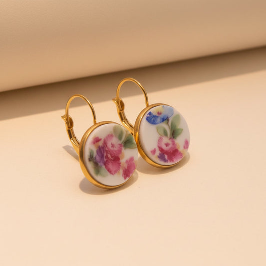Nahaufnahme von zwei runden Upcycling-Ohrhängern aus weißem Porzellan mit einem reichhaltigen, bunten Floribunda-Blumenstrauß-Motiv in Pink, Blau und Lila, eingefasst in goldfarbenem Edelstahl auf beigem Hintergrund. Die Ohrringe sind leicht seitlich von oben fotografiert.