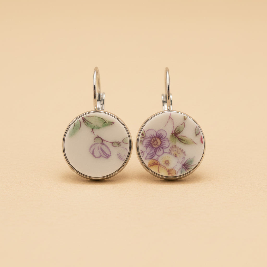 Zwei handgefertigte Porzellan-Ohrhänger des Modells Lilaura von Edelatelier in silberfarbenen Edelstahlfassungen. Die runden Inlays zeigen filigrane florale Motive aus Upcycling-Porzellan: links eine violette Blütenranke und rechts ein detailliertes Bukett mit lila und gelben Blumen. Das Paar ist auf einem neutralen, beigefarbenen Hintergrund vor einem sanften Schattenwurf abgebildet.