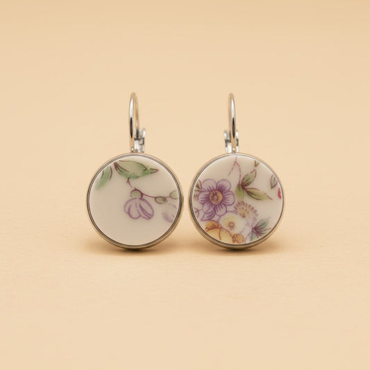 Zwei handgefertigte Porzellan-Ohrhänger des Modells Lilaura von Edelatelier in silberfarbenen Edelstahlfassungen. Die runden Inlays zeigen filigrane florale Motive aus Upcycling-Porzellan: links eine violette Blütenranke und rechts ein detailliertes Bukett mit lila und gelben Blumen. Das Paar ist auf einem neutralen, beigefarbenen Hintergrund vor einem sanften Schattenwurf abgebildet.