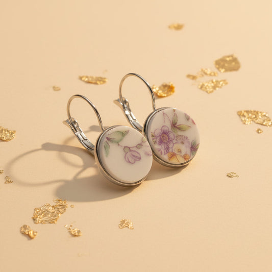 Seitenansicht der Lilaura Porzellan-Ohrringe von Edelatelier, die die stabilen Klappbrisuren aus silberfarbenem Edelstahl zeigen. Die handgeschliffenen Porzellan-Inlays mit ihren violetten und gelben Blütenmotiven sind in der Fassung deutlich erkennbar. Die Ohrhänger liegen auf einer beigen Fläche, dekoriert mit feinen Goldflocken.
