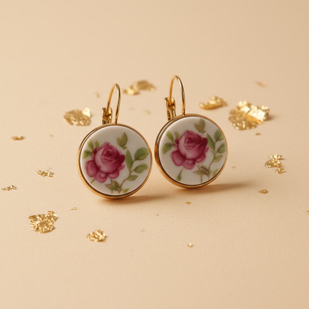 Nahaufnahme von zwei handgefertigten Upcycling-Ohrhängern aus weißem Porzellan mit einem detaillierten Rosen-Motiv in kräftigem Pink und grünen Blättern, gefasst in goldfarbenen Edelstahl auf einem beigen Untergrund mit Goldflocken.