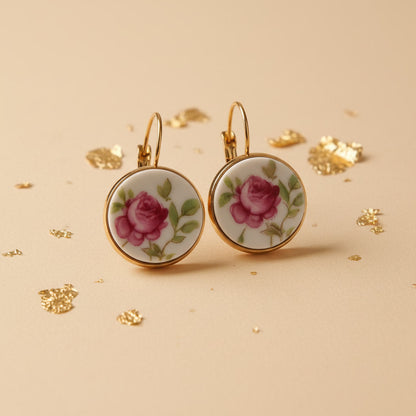 Nahaufnahme von zwei handgefertigten Upcycling-Ohrhängern aus weißem Porzellan mit einem detaillierten Rosen-Motiv in kräftigem Pink und grünen Blättern, gefasst in goldfarbenen Edelstahl auf einem beigen Untergrund mit Goldflocken.