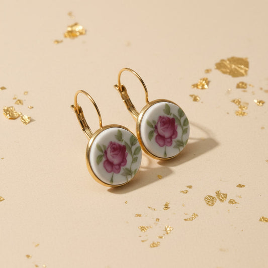 Ein Paar runde Upcycling-Ohrhänger, seitlich fotografiert, aus weißem Porzellan mit einem detaillierten Rosen-Motiv in leuchtendem Pink und Grün, eingefasst in goldfarbenen Edelstahl auf einem beigen Untergrund mit dekorativen Goldflocken.