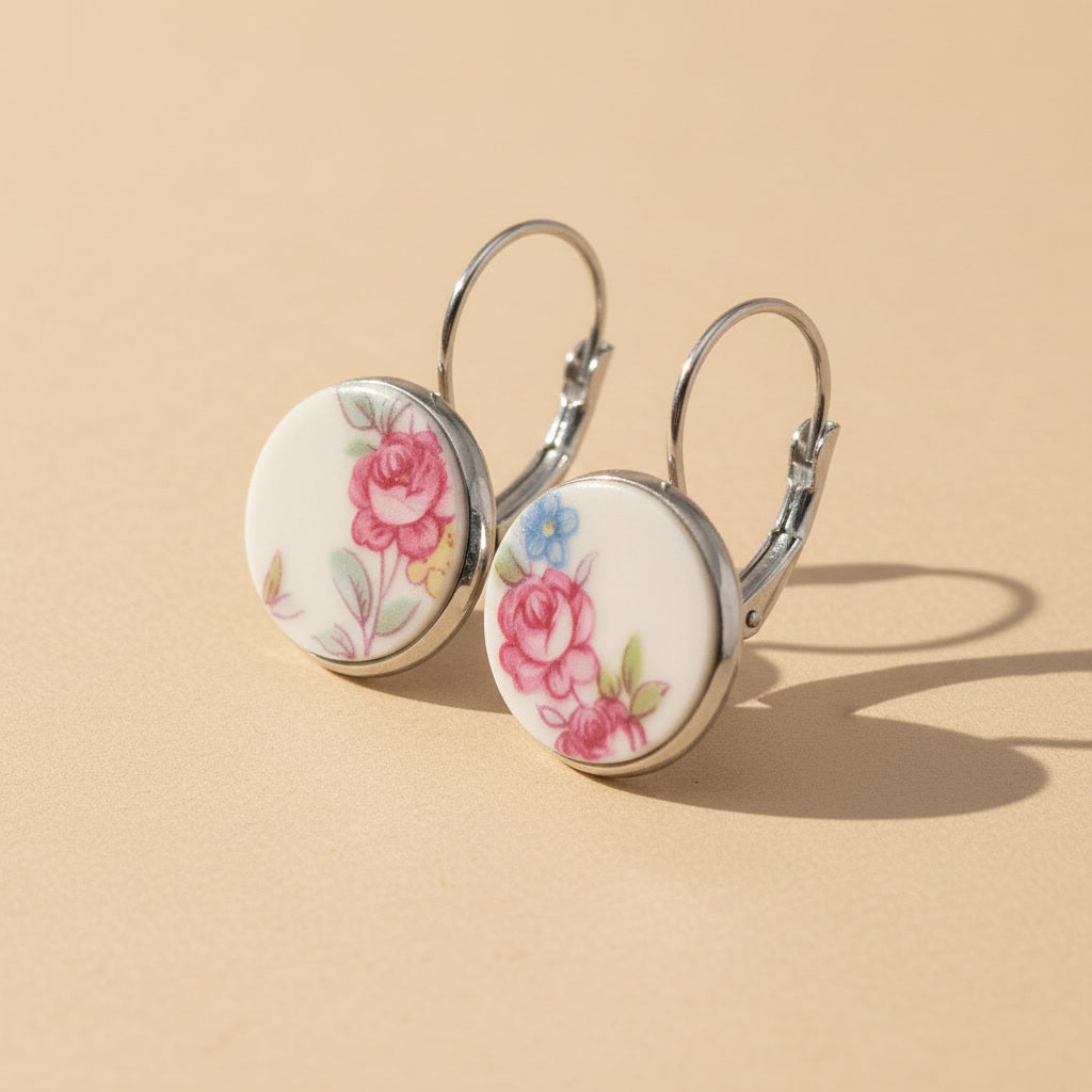 Seitenansicht der Rosula Porzellan-Ohrringe von Edelatelier, welche die hochwertigen Klappbrisuren aus Edelstahl zeigen. Die runden Inlays aus Upcycling-Porzellan präsentieren verschiedene Stadien von Rosenmotiven in Pink und Rosé mit feinen blauen Farbtupfern. Das Paar ist auf einem beigen Hintergrund mit sanfter Schattenbildung arrangiert.