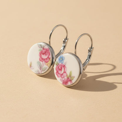 Seitenansicht der Rosula Porzellan-Ohrringe von Edelatelier, welche die hochwertigen Klappbrisuren aus Edelstahl zeigen. Die runden Inlays aus Upcycling-Porzellan präsentieren verschiedene Stadien von Rosenmotiven in Pink und Rosé mit feinen blauen Farbtupfern. Das Paar ist auf einem beigen Hintergrund mit sanfter Schattenbildung arrangiert.