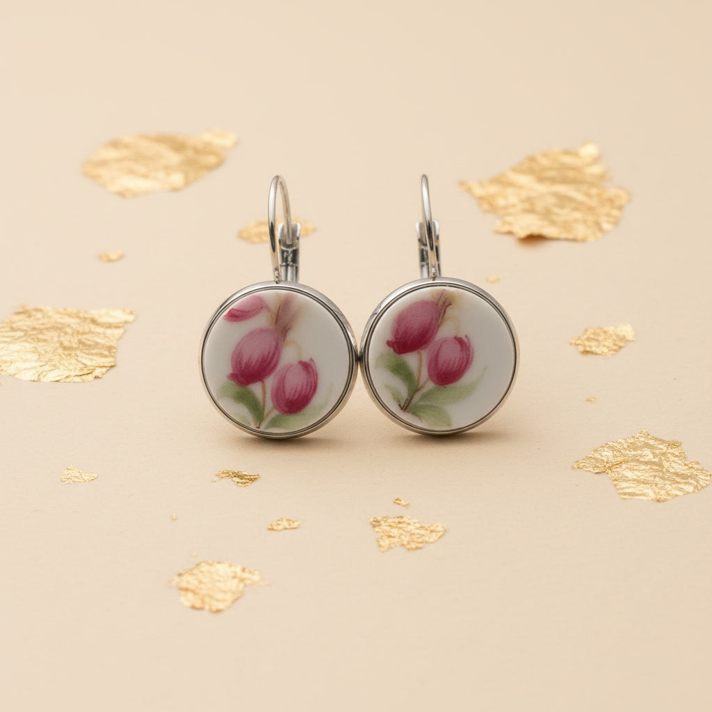 Nahaufnahme von zwei handgefertigten Upcycling-Ohrhängern aus weißem Porzellan, frontal fotografiert, mit einem filigranen Blumenmotiv in dunklem Pink und Grün, gefasst in silberfarbenen Edelstahl auf einem beigefarbenen Hintergrund mit dekorativen Goldflocken.