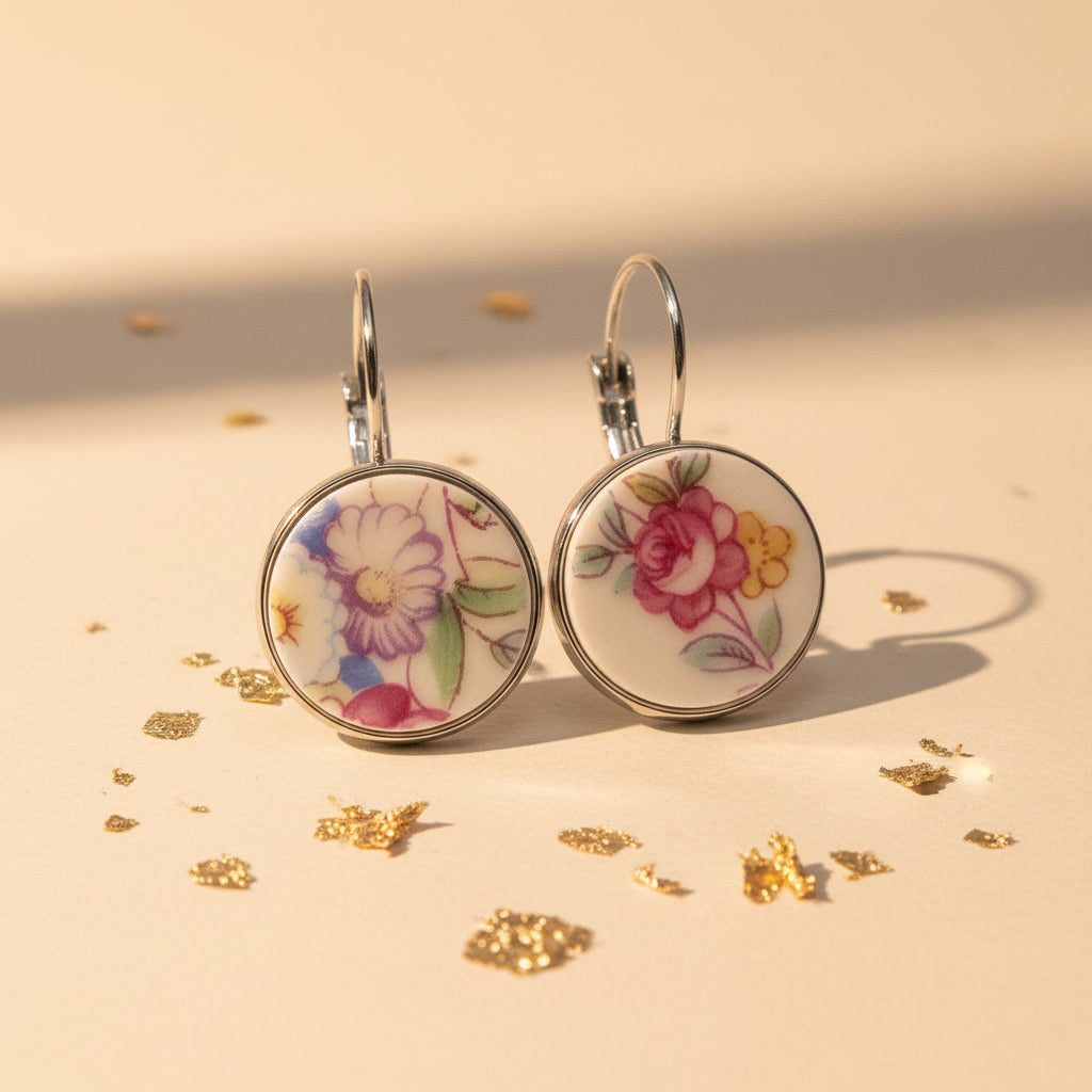 Zwei handgefertigte Porzellan-Ohrhänger des Modells Varietas von Edelatelier in silberfarbenen Edelstahlfassungen. Die runden Inlays zeigen unterschiedliche florale Motive aus Upcycling-Porzellan: links eine violett-weiße Blüte mit grünen Blättern und rechts eine pinkfarbene Rose mit gelben Akzenten. Die Ohrringe liegen dekorativ auf einem beigefarbenen Hintergrund, umgeben von feinen Goldflocken unter warmem Studio-Licht.