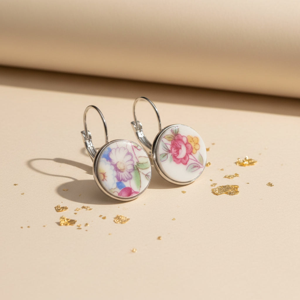 Seitenansicht der Varietas Porzellan-Ohrringe von Edelatelier, die die hochwertigen Klappbrisuren aus silberfarbenem Edelstahl zeigen. Die flachen, handgeschliffenen Porzellan-Inlays mit ihren farbenfrohen Blumenmustern sind deutlich erkennbar. Die Ohrhänger stehen auf einer beigefarbenen Fläche mit Goldakzenten, was das leichte und elegante Design unterstreicht.