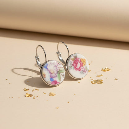 Seitenansicht der Varietas Porzellan-Ohrringe von Edelatelier, die die hochwertigen Klappbrisuren aus silberfarbenem Edelstahl zeigen. Die flachen, handgeschliffenen Porzellan-Inlays mit ihren farbenfrohen Blumenmustern sind deutlich erkennbar. Die Ohrhänger stehen auf einer beigefarbenen Fläche mit Goldakzenten, was das leichte und elegante Design unterstreicht.