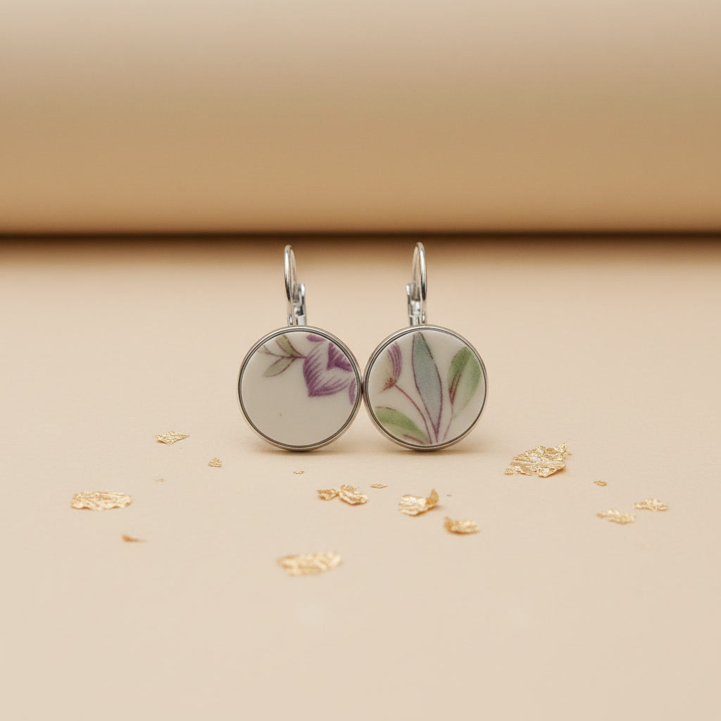 Zwei handgefertigte Porzellan-Ohrhänger des Modells Viola von Edelatelier in silberfarbenen Edelstahlfassungen. Die runden Inlays aus Upcycling-Porzellan zeigen ein dezentes florales Muster mit zart-violetten Blütenkelchen und schmalen, salbeigrünen Blättern. Das Paar ist auf einem beigefarbenen Hintergrund mit dekorativen Goldflocken und sanfter Schattenbildung arrangiert.