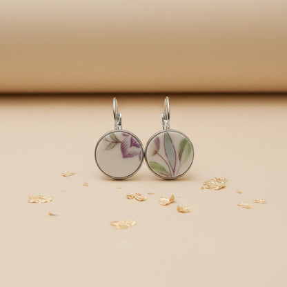 Zwei handgefertigte Porzellan-Ohrhänger des Modells Viola von Edelatelier in silberfarbenen Edelstahlfassungen. Die runden Inlays aus Upcycling-Porzellan zeigen ein dezentes florales Muster mit zart-violetten Blütenkelchen und schmalen, salbeigrünen Blättern. Das Paar ist auf einem beigefarbenen Hintergrund mit dekorativen Goldflocken und sanfter Schattenbildung arrangiert.